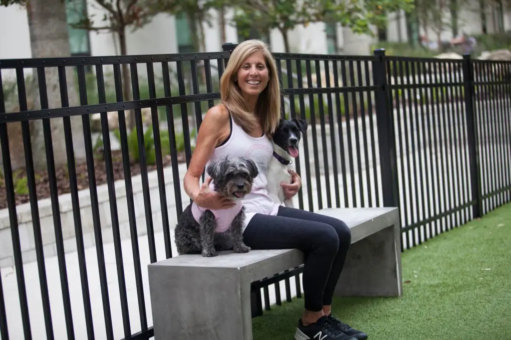 Marilyn Moss, owner of Functional Fitness Guru, sits on a bench in Tampa, smiling and holding two small dogs. She is dressed in workout attire.